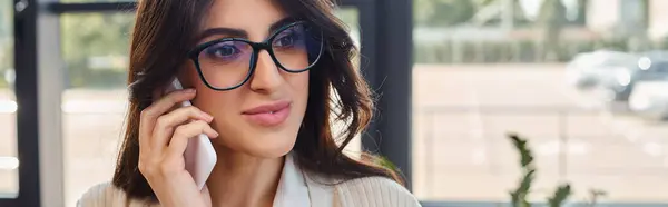 A professional businesswoman wearing glasses conducts a phone call in a modern office setting near her workspace. — Stock Photo