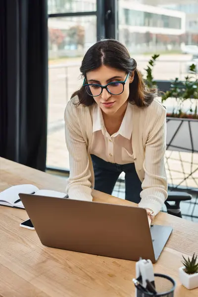 Una donna d'affari con gli occhiali si concentra sul suo computer portatile, gestendo un franchising in un moderno ufficio. — Foto stock