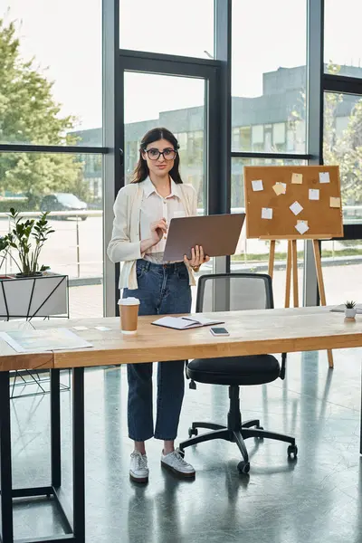 A businesswoman confidently stands by her modern office table, focused on her laptop, symbolizing the franchise concept. — Stock Photo