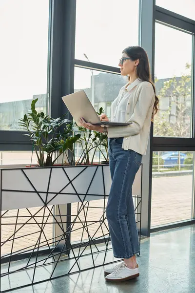 Una mujer de negocios está al lado de una ventana, enfocada en su computadora portátil en un espacio de oficina moderno con un concepto de franquicia. - foto de stock