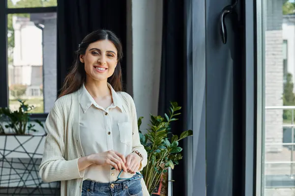 A businesswoman stands confidently in front of a window, in a modern office setting near her workspace. — Stock Photo
