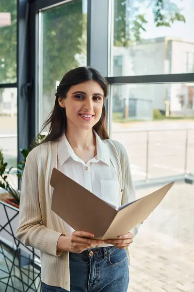 A businesswoman stands confidently in front of a window, holding a folder, embodying the essence of a modern CEO in a franchise office. — Stock Photo