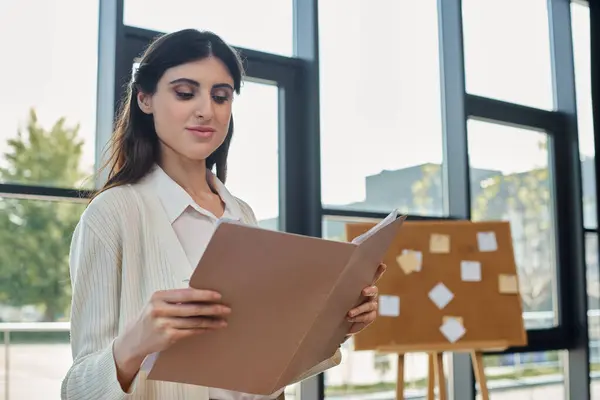 A businesswoman stands near her workspace, holding a paper in front of a window, contemplating her next steps in the franchise world. — Stock Photo