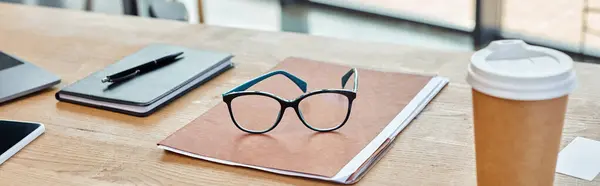 Un par de gafas descansa en un vibrante cuaderno junto a una taza de café humeante, ubicado en un moderno espacio de trabajo de oficina. - foto de stock