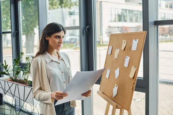 Una mujer de negocios está parada frente a una tabla, sosteniendo un pedazo de papel en una oficina moderna, encarnando el concepto de franquicia. - foto de stock