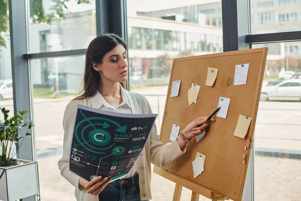 Uma mulher de negócios fica na frente do conselho, segurando gráficos em um ambiente de escritório moderno. — Fotografia de Stock