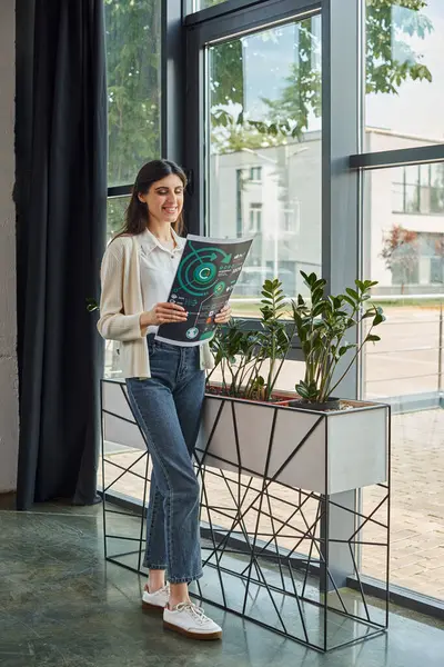 A modern businesswoman stands near her workspace, holding charts, gazing out of a window. — Stock Photo