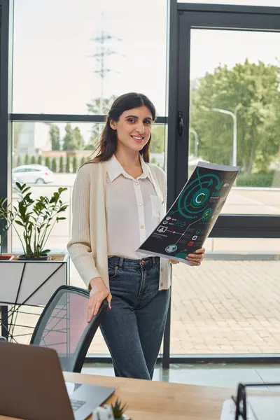 A modern businesswoman exudes knowledge and sophistication as she stands in her office, holding charts with confidence. — Stock Photo