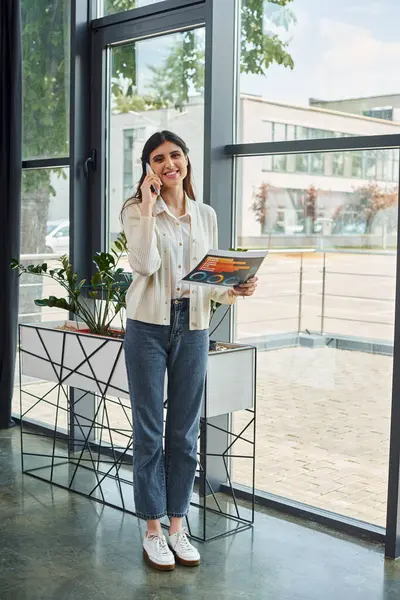 Una donna d'affari in piedi vicino a una finestra in un ufficio moderno, impegnata in una telefonata, in mostra un concetto di franchising. — Foto stock