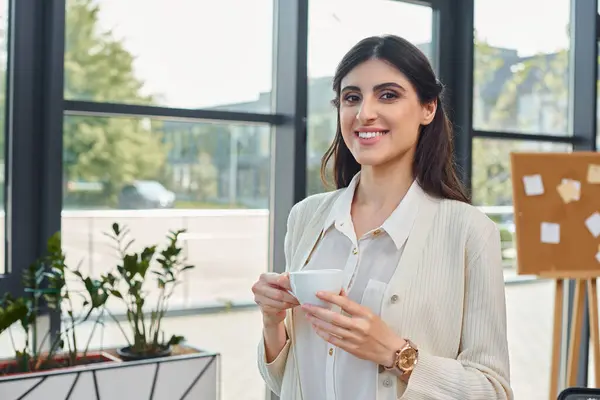 A businesswoman savors a cup of coffee against a cityscape backdrop through a large window in a modern office. — Stock Photo