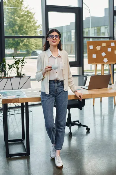 Una mujer de negocios está de pie con confianza junto a una mesa con una computadora portátil en un entorno de oficina moderno, encarnando el concepto de franquicia. - foto de stock