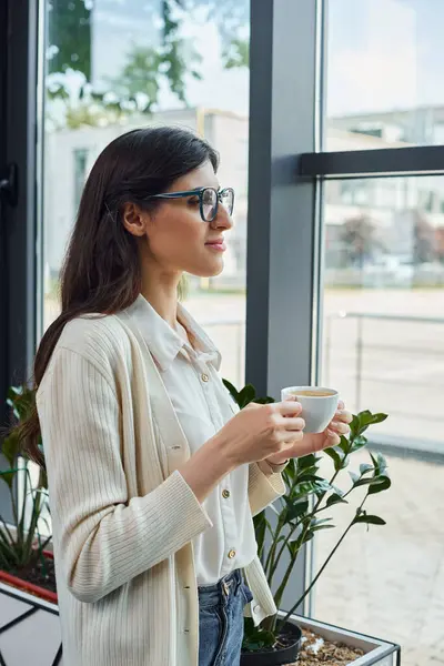 A modern businesswoman holds a cup, standing in front of a window in an office, deep in thought. — Stock Photo