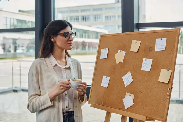A businesswoman stands in front of a board, holding a cup of coffee in a modern office setting. — Stock Photo