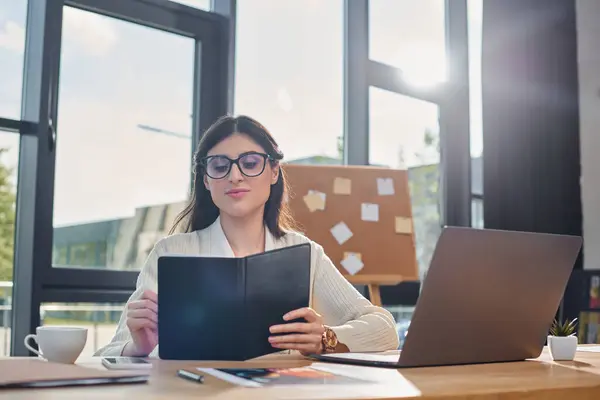Una donna d'affari si siede in un ufficio moderno su una scrivania, focalizzata sul suo computer portatile, incarnando il concetto di franchising.. — Foto stock