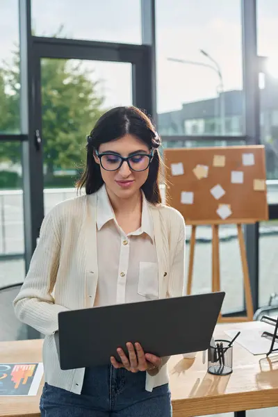Una mujer de negocios se para con confianza frente a una computadora portátil en un entorno de oficina moderno, encarnando el concepto de franquicia. - foto de stock
