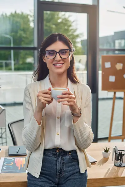 Una empresaria moderna en gafas saborea una taza de café en su espacio de trabajo de oficina, encarnando la esencia de un concepto de franquicia próspero. - foto de stock