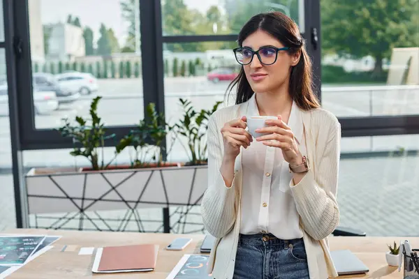 Une femme d'affaires sophistiquée dans des lunettes chérit une tasse chaude de café dans un bureau moderne, incarnant un concept de franchise. — Photo de stock