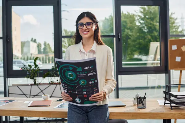 Una mujer de negocios está con confianza en una oficina moderna, sosteniendo gráficos cerca de su espacio de trabajo en un entorno de concepto de franquicia. - foto de stock
