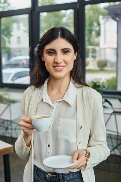 Una mujer de negocios en un moderno entorno de oficina sostiene con gracia una taza de café y un plato, tomando un descanso de su trabajo. - foto de stock