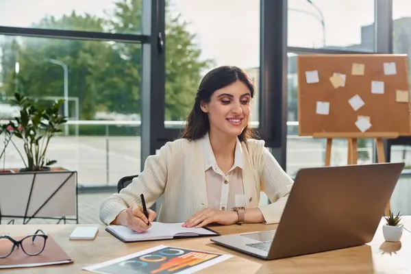 Una mujer de negocios se sienta en una mesa en una oficina moderna, enfocada en la pantalla de su computadora portátil mientras profundiza en el concepto de franquicia. - foto de stock