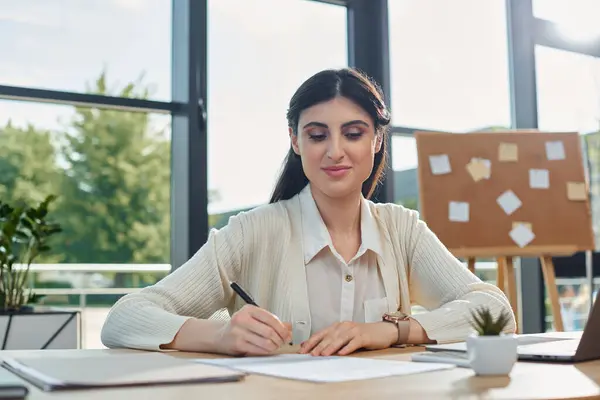 Eine Geschäftsfrau in einem modernen Büro sitzt an einem Schreibtisch und konzentriert sich darauf, auf ein Blatt Papier zu schreiben. — Stockfoto