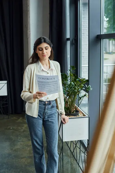 Eine moderne Geschäftsfrau liest einen Vertrag, während sie an einem Fenster in ihrem Büro steht, umgeben von einem Franchise-Arbeitsplatz. — Stockfoto
