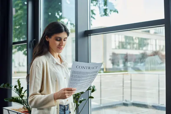 Una empresaria moderna lee un contrato estando de pie junto a una ventana en su oficina, rodeada por un espacio de trabajo de franquicia. - foto de stock
