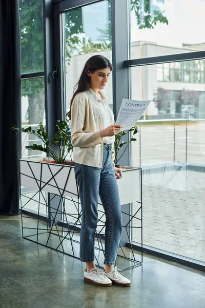 Eine Geschäftsfrau steht vor einem Fenster und hält einen Vertrag in einem modernen Büroraum, tief in Gedanken. — Stockfoto