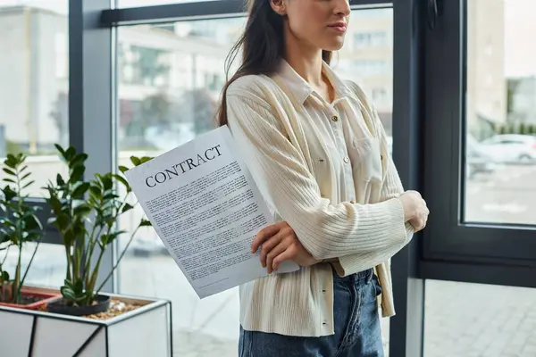 Une femme d'affaires moderne se tient près d'une fenêtre, tenant un morceau de papier, envisageant les prochaines étapes de son concept de franchise. — Photo de stock