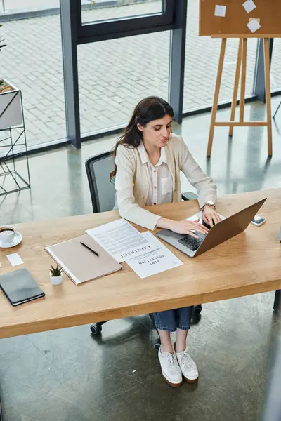 Una empresaria en una oficina moderna, centrada en su portátil en una mesa, inmersa en el mundo del emprendimiento. - foto de stock