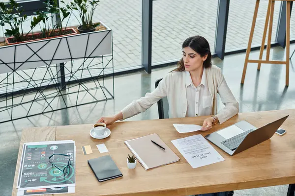 Una mujer de negocios se sienta en su escritorio rodeada de una computadora portátil y papeles, centrada en su trabajo de franquicia en una oficina moderna. - foto de stock