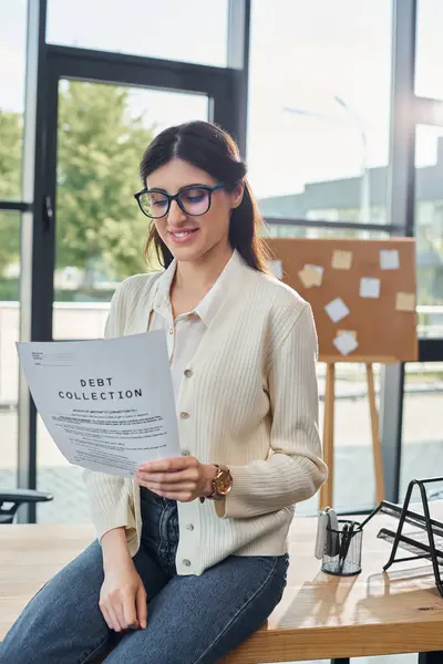 Бізнес-леді, сидячи на столі, впевнено тримає аркуш паперу в сучасному офісному приміщенні біля її робочого простору. — стокове фото