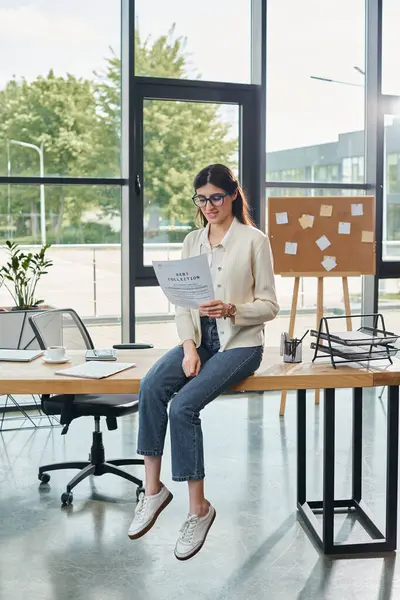 A modern businesswoman sits on a table, engrossed in reading a piece of paper in a modern office setting near her workspace. — Stock Photo