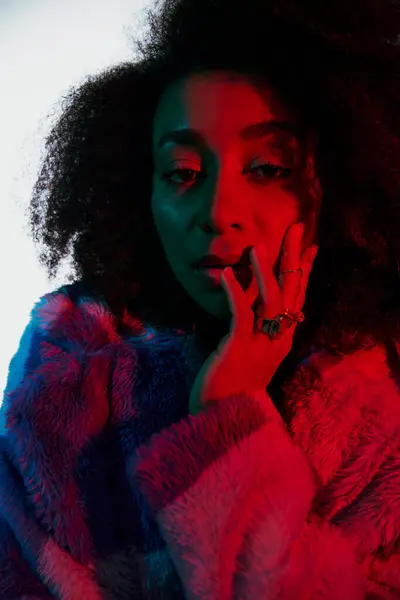 Elegant african american woman in faux fur with curly hair looking at camera in red and blue lights — Stock Photo