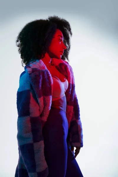 Seductora mujer afroamericana en piel sintética mirando hacia otro lado en luces rojas y azules, concepto de moda - foto de stock