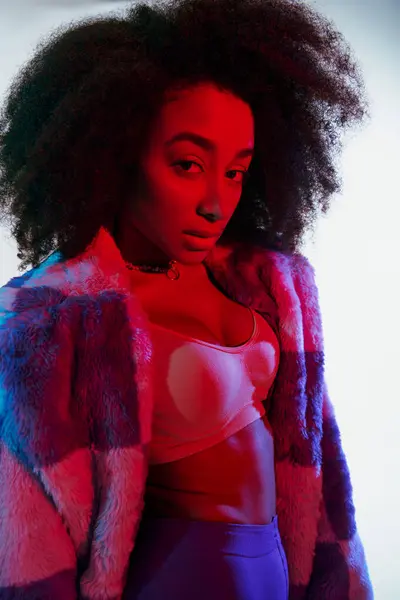 Debonair mulher americana africana em peles falsas com cabelo encaracolado olhando para a câmera em luzes vermelhas e azuis — Fotografia de Stock