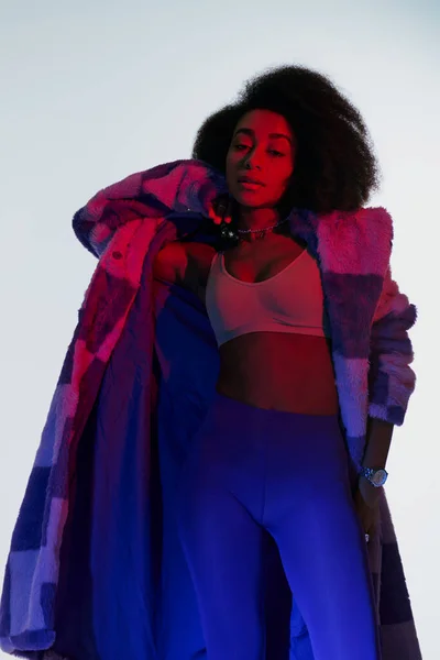 Sexy donna afroamericana in reggiseno e pelliccia sintetica guardando la fotocamera circondata da luci rosse e blu — Foto stock