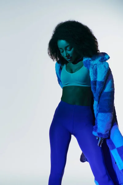 Seductora mujer afroamericana con estilo en piel sintética y sujetador posando en luces azules, concepto de moda - foto de stock