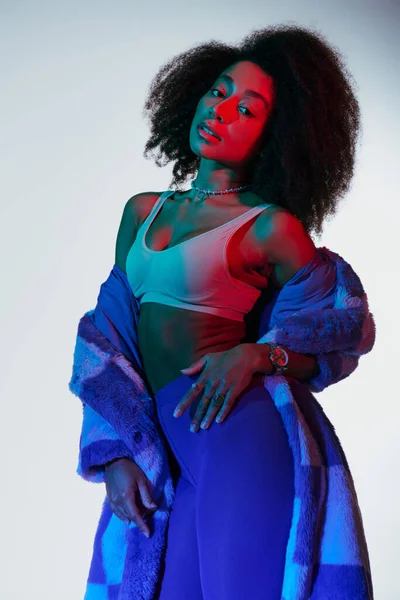 Atractiva mujer afroamericana sexy en piel sintética con estilo de moda mirando a la cámara en luces - foto de stock