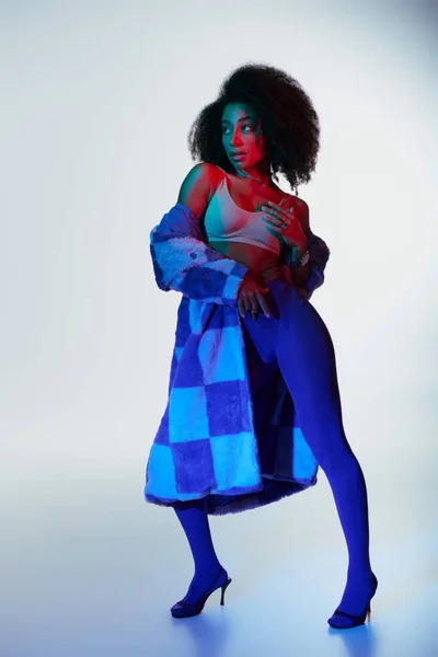 Tentador modelo femenino afroamericano con estilo en piel sintética de moda buscando camino en las luces - foto de stock