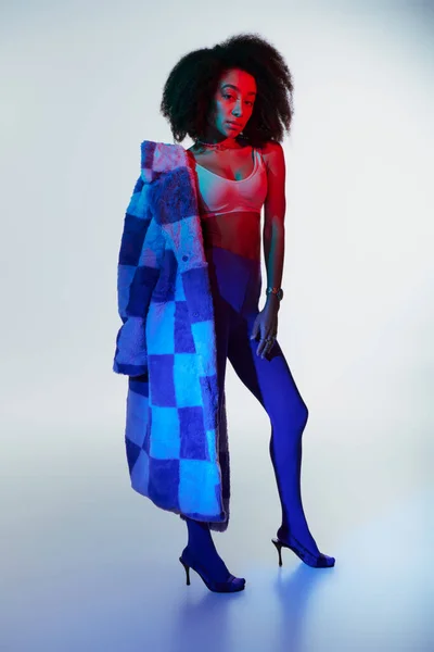 Hermosa mujer afroamericana sexy en piel sintética con estilo de moda mirando a la cámara en luces - foto de stock