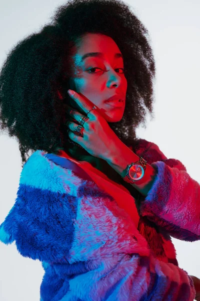Atraente mulher americana africana na moda em peles falsas olhando para a câmera em luzes vermelhas e azuis — Fotografia de Stock