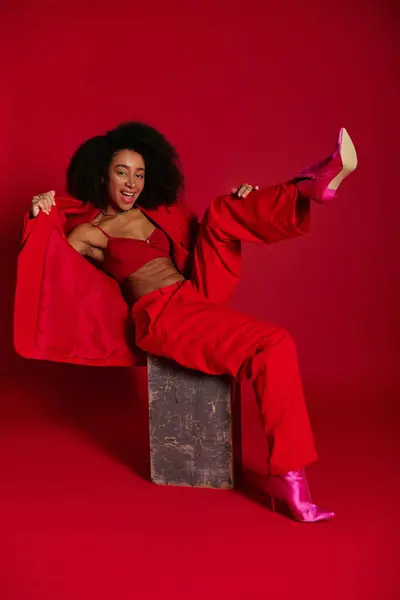 Mulher americana africana alegre em traje chique vermelho sentado e sorrindo para a câmera, conceito de moda — Fotografia de Stock
