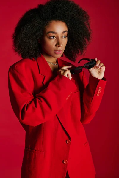 Atractiva mujer afroamericana en traje vibrante con gafas de sol mirando hacia otro lado, concepto de moda - foto de stock