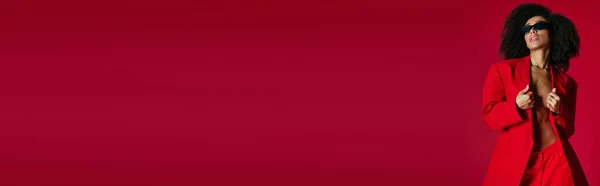 Alluring african american woman in red unbuttoned blazer with sunglasses on red backdrop, banner — Stock Photo