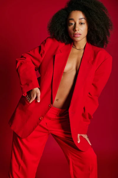 Seductora mujer afroamericana en chaqueta mirando a la cámara sobre fondo rojo, concepto de moda - foto de stock