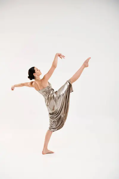A graceful young woman in a long and shiny silver dress elegantly dances in a studio setting against a white background. — Stock Photo