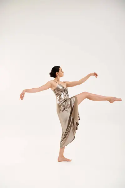 A young woman exudes grace and elegance as she dances in a long, shiny silver dress in a studio setting against a white backdrop. — Stock Photo