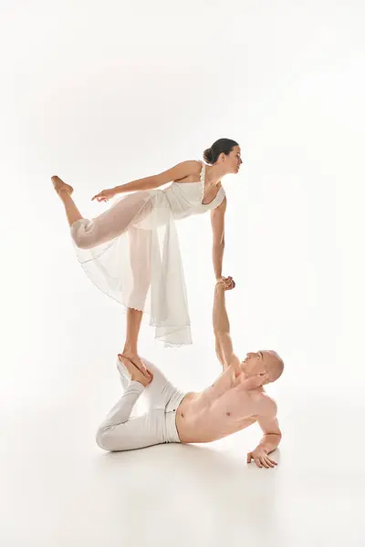 A shirtless young man and a woman in a white dress perform acrobatic exercises together in a studio setting on a white background. — Stock Photo
