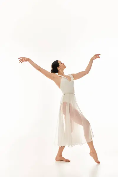 A graceful young woman dances in a flowing white dress against a white studio backdrop. — Stock Photo